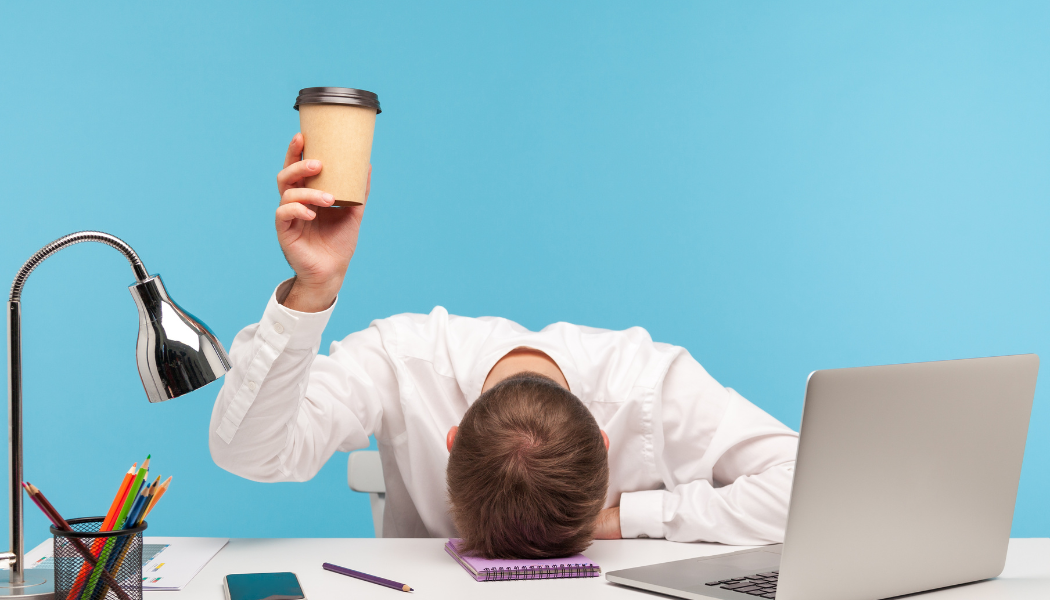 Mann am Schreibtisch hält Kaffeebecher hoch, wirkt erschöpft – Symbolbild für Müdigkeit im Alltag